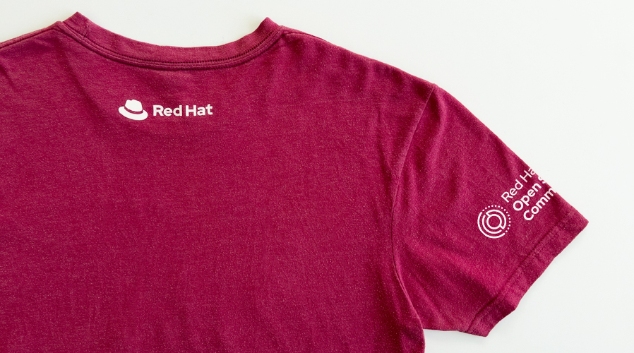 Photography of a t-shirt with the Red Hat Open Studio Community inititiave logo on the sleeve and the Red Hat logo on the neck area.